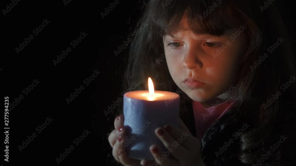 Sad child looks at the flame of a candle. A sad little girl looks at a burning candle. Sad child looks at the flame of a candle. A sad little girl looks at a burning candle.