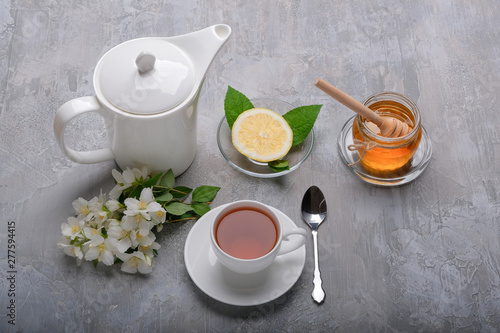 A cup of tea, a kettle, a lemon, a jar of honey and jasmine flowers on a grey background.