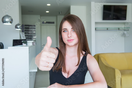 Close up Portrait of happy smiling young woman with deep decollete and thumb up hand sitting behind the desk in the office hall. Selective focus, copy space.