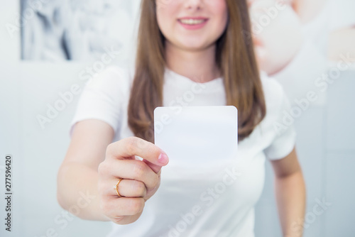 Business card or gift card. Blurred Happy and excited caucasian woman in casual clothes showing focused blank empty paper card with copy space for your design on light indoor background.