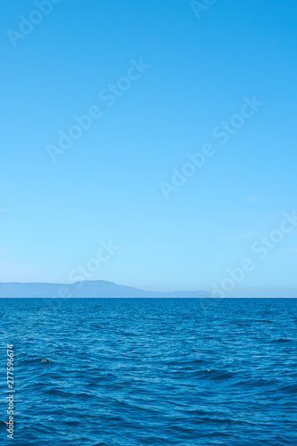 Fototapeta Naklejka Na Ścianę i Meble -  Blue sea water with foggy rocky island on a background under a clear blue sky. Copy space. 