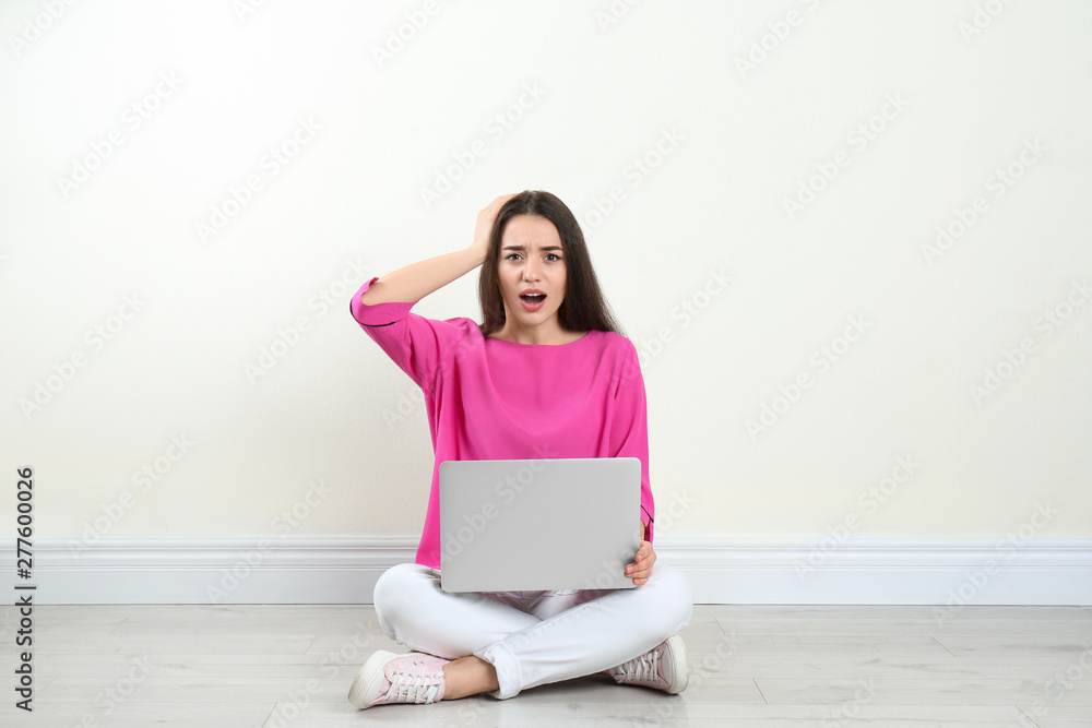 Shocked young woman with laptop sitting on floor near light wall indoors Shocked young woman with laptop sitting on floor near light wall indoors