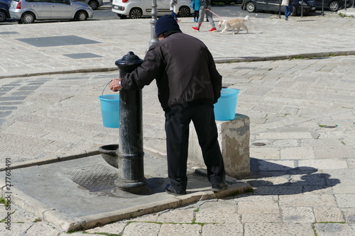 Anziano signore in piazza alla fontana mentre riempie d'acqua due secchi di plastica photo