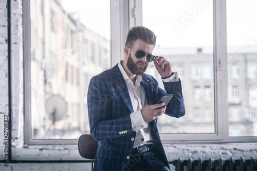 He will melt your heart. Handsome young bearded man in full suit and eyewear looking to his cell phone while sitting on the stool