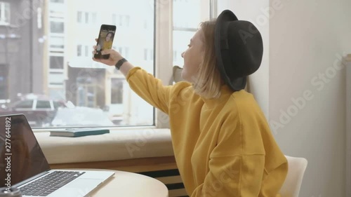 Young pretty blonde female blogger using smart phone, typing, taking selfie, chatting, while sitting at the cafe, using laptop on the table, near the window, she is wearing yellow sweater and hat photo