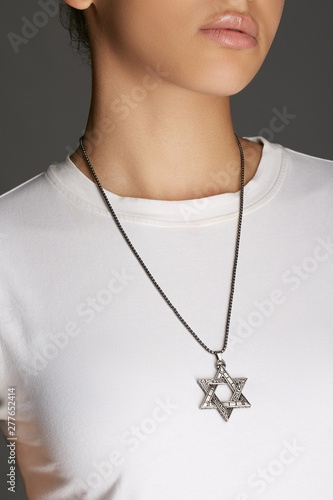 Cropped top and bottom photo of a woman with a necklace on her neck. The model on a gray background is dressed in a white tee-shirt. You can see Star of David pendant with engraved intricate patterns 