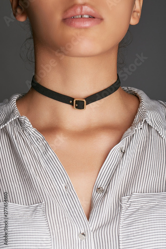 Cropped top and bottom photo of a woman with a choker necklace on her neck. The model on a gray background is dressed in a white tee-shirt. Leather choker necklace is decorated with a metal buckle at 