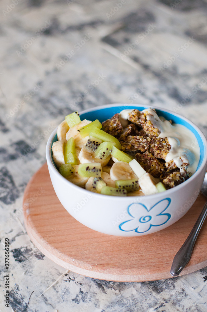 Greek yogurt, granola, kiwi and banana in bowl with a spoon on a gray concrete background, wooden tray, vertical format. Fitness diet for weight loss, proper and tasty food