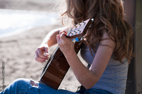 Cute young woman wearing long blond hair playing guitar sitting on a beach