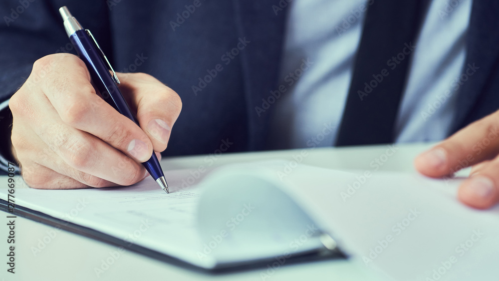 Hand of businessman in suit filling and signing with blue pen partnership agreement form clipped to pad closeup. Hand of businessman in suit filling and signing with blue pen partnership agreement form clipped to pad closeup.