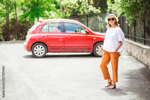 Car rental trip. Stylish adult woman standing and smiling by the small red car to drive, rent, travel, buy it. © Anna