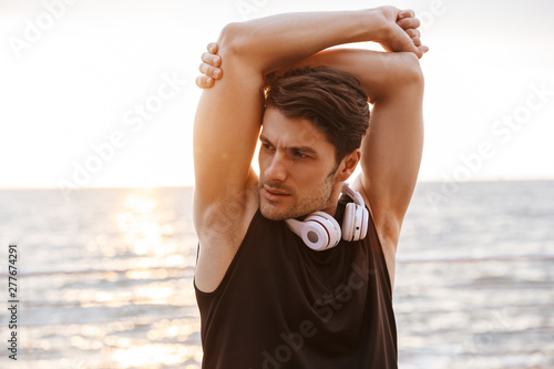Photo unshaven man in tracksuit with headphones doing exercise while working out at seaside