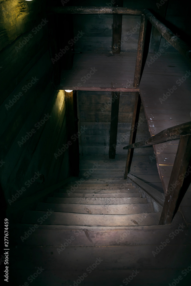 Darkness and horror in a ghostly building. Inside on a dark wooden staircase with steps down to the basement of an old abandoned house with black walls mystical light from a dim lamp on the floor