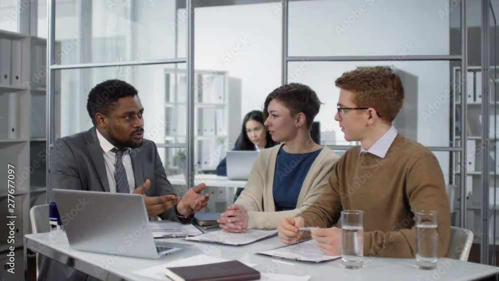Full shot of Afro-American real estate agent consulting young Caucasian couple about house lease, demonstrating available properties on laptop and discussing contract terms and conditions