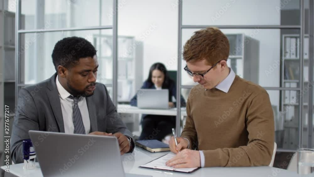 Waist-up shot of 30-something Caucasian man signing contract in African American solicitor’s office, who is then stamping it with seal, while female staff are working on laptops in blurred background Waist-up shot of 30-something Caucasian man signing contract in African American solicitor’s office, who is then stamping it with seal, while female staff are working on laptops in blurred background