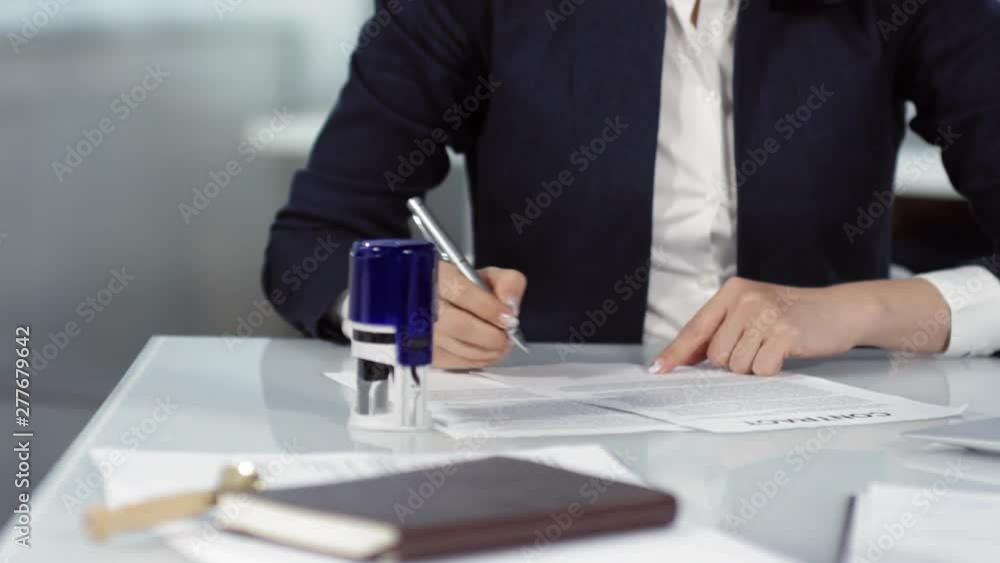 Tracking hands and body shot of business woman, dressed in white blouse and jacket, sitting at desk, reading through contract, signing it and sealing with self-inking rubber stamp