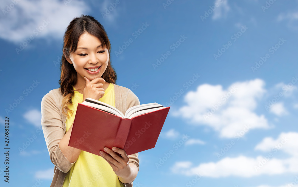 people, ethnicity and portrait concept - happy asian young woman reading book over blue sky and clouds background people, ethnicity and portrait concept - happy asian young woman reading book over blue sky and clouds background