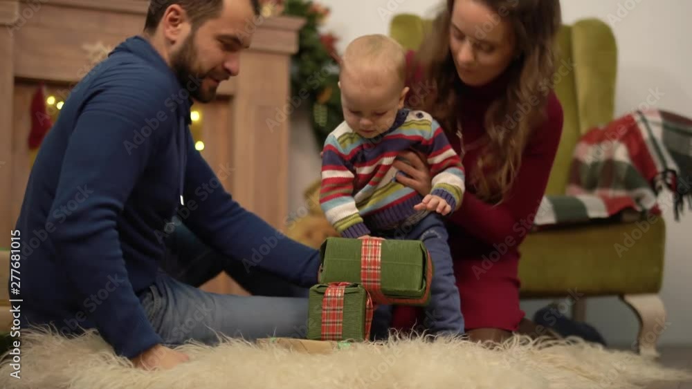 Mother, father and little baby sitting on the floor in the room with christmas decoration. Man gives small present box to child crawling on fluffy carpet. Happy family celebrating Christmas together Mother, father and little baby sitting on the floor in the room with christmas decoration. Man gives small present box to child crawling on fluffy carpet. Happy family celebrating Christmas together