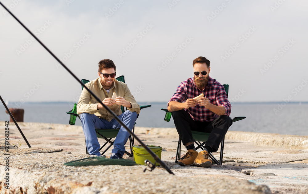 leisure and people concept - happy friends fishing and eating sandwiches on pier at sea