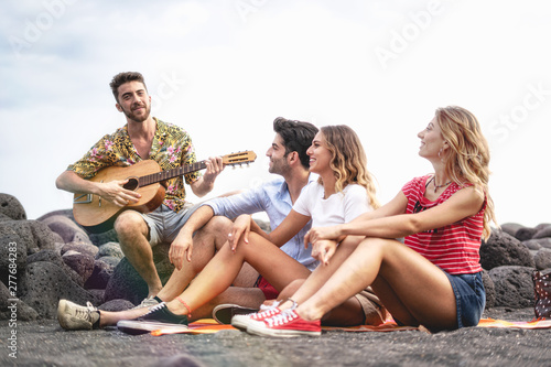 Group of young friends having good time at the beach playing guitar and singing songs at sunset. Youth lifestyle concept