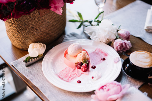 Delicious fresh peony dessert at the restaurant: cream cheese with peony flavoured ice cream on a white plate, a cup of cappuccino coffee and a wicker basket full of blossoming peonies on the table