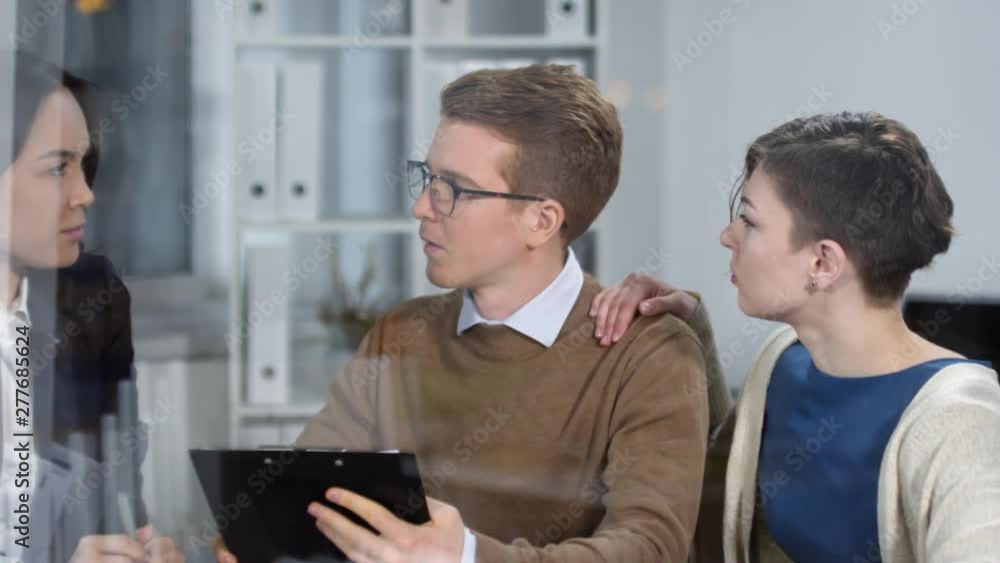 Hidden camera tracking shot of young Caucasian man and woman sitting in solicitors office, holding clipboard with document draft and discussing it with professionally dressed Asian female legal expert Hidden camera tracking shot of young Caucasian man and woman sitting in solicitors office, holding clipboard with document draft and discussing it with professionally dressed Asian female legal expert