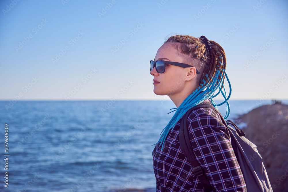 Girl with blue dreadlocks and dark glasses against the sea, illuminated by the evening sun.