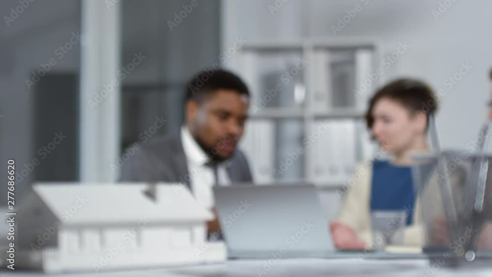Tracking rack focus shot of white scaled house model in foreground, and Afro-American real estate broker, wearing business suit and tie, engaged in emotional argument with Caucasian client couple Tracking rack focus shot of white scaled house model in foreground, and Afro-American real estate broker, wearing business suit and tie, engaged in emotional argument with Caucasian client couple