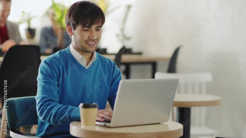 Smiling young Middle Eastern man sitting at table in cafeteria and working on laptop computer, tracking right medium shot Smiling young Middle Eastern man sitting at table in cafeteria and working on laptop computer, tracking right medium shot