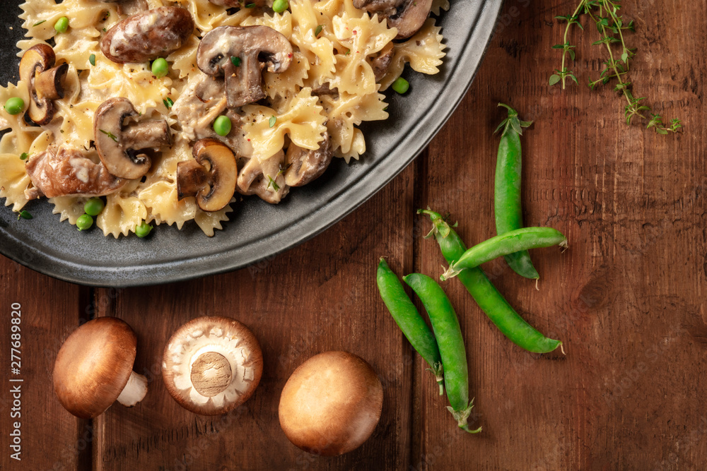 Fototapeta premium Mushroom and cheese pasta. Farfalle with cremini and green peas, shot from the top with ingredients on a dark rustic wooden background with a place for text
