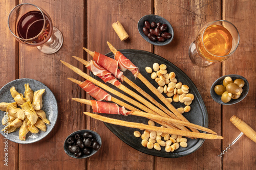 Italian antipasti. Grissini breadsticks with parma ham and roasted almonds, with olives and artichokes, shot from the top on a dark rustic wooden background with wine glasses