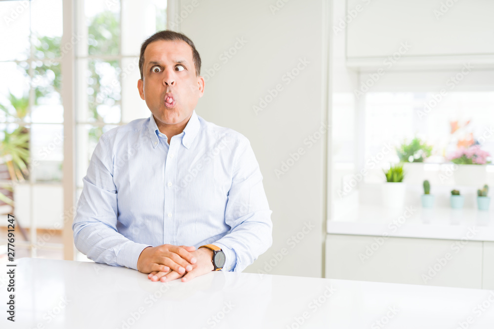 Middle age man sitting at home making fish face with lips, crazy and comical gesture. Funny expression.