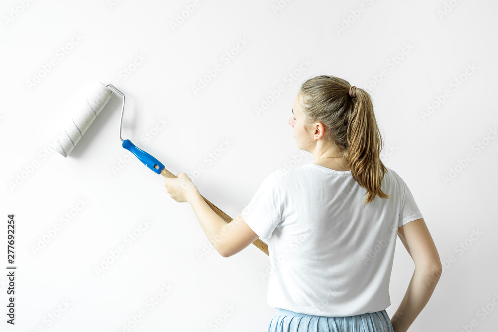 Young happy woman painting interior wall with paint roller in new house. A young girl makes repairs: paints the walls with white paint using a roller. Young happy woman painting interior wall with paint roller in new house. A young girl makes repairs: paints the walls with white paint using a roller.
