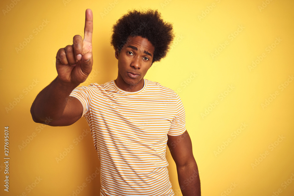 American man with afro hair wearing striped t-shirt standing over isolated yellow background Pointing with finger up and angry expression, showing no gesture American man with afro hair wearing striped t-shirt standing over isolated yellow background Pointing with finger up and angry expression, showing no gesture