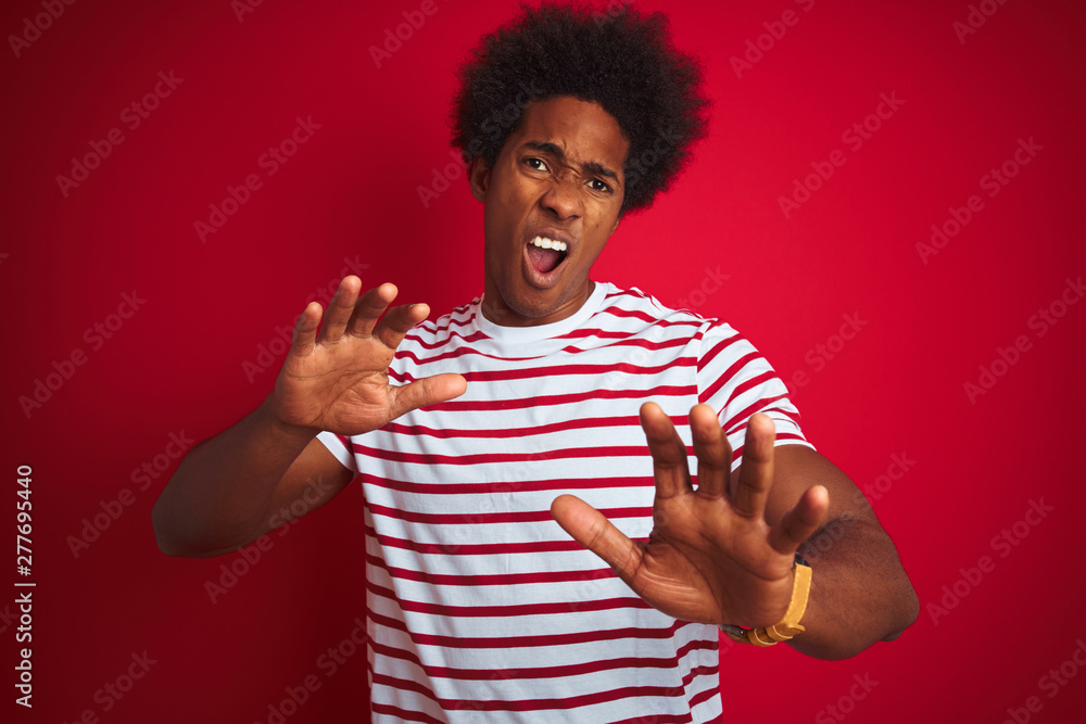 Young african american man with afro hair wearing striped t-shirt over isolated red background afraid and terrified with fear expression stop gesture with hands, shouting in shock. Panic concept. Young african american man with afro hair wearing striped t-shirt over isolated red background afraid and terrified with fear expression stop gesture with hands, shouting in shock. Panic concept.