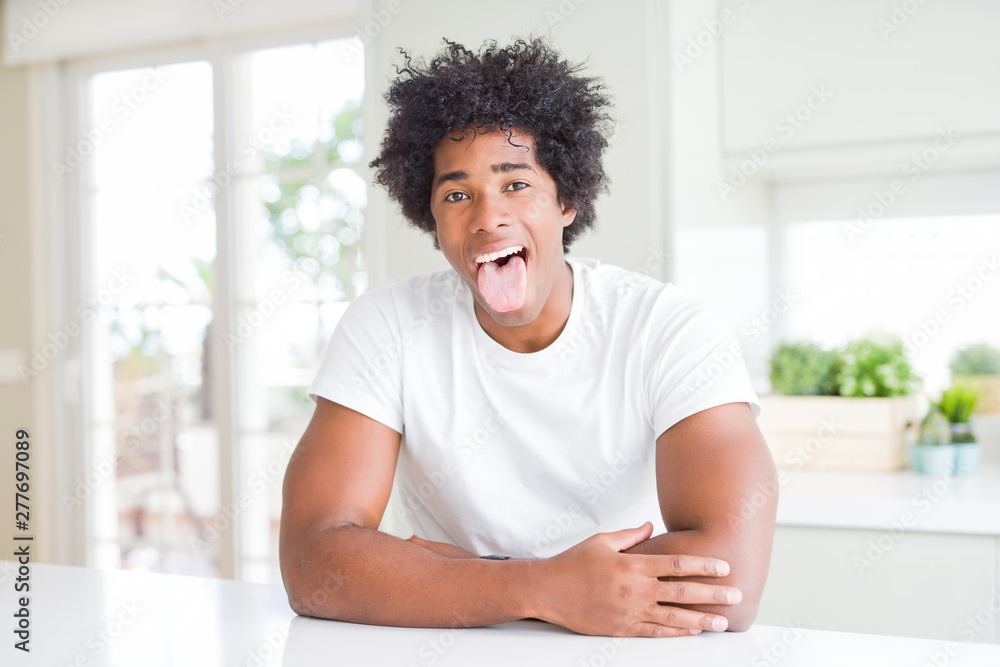 Young african american man wearing casual white t-shirt sitting at home sticking tongue out happy with funny expression. Emotion concept. Young african american man wearing casual white t-shirt sitting at home sticking tongue out happy with funny expression. Emotion concept.
