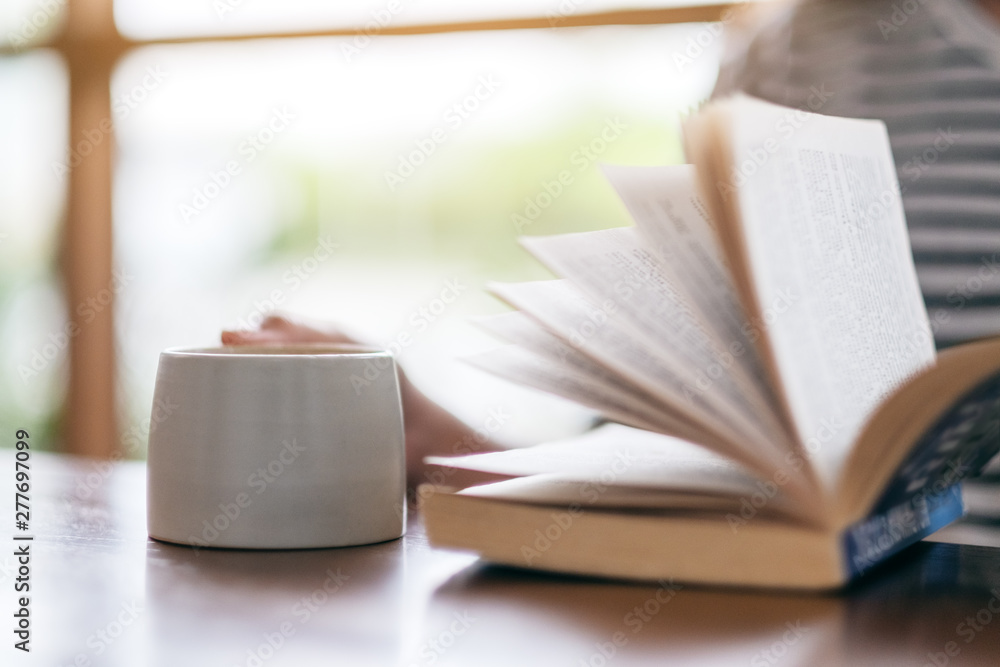 Fototapeta premium Closeup image of a woman holding and reading a book while drinking coffee on wooden table