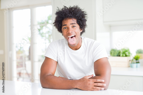 Young african american man wearing casual white t-shirt sitting at home sticking tongue out happy with funny expression. Emotion concept.