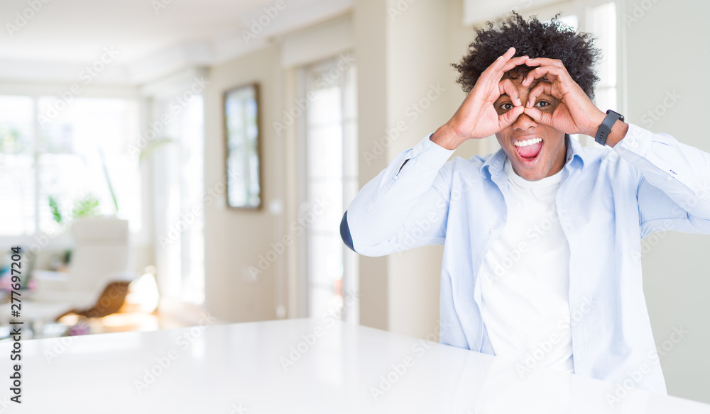 African American man at home doing ok gesture like binoculars sticking tongue out, eyes looking through fingers. Crazy expression.