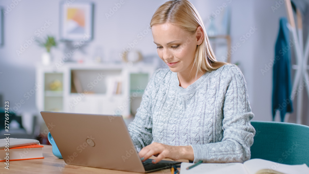 Beautiful Blonde Woman Works on a Laptop while Sitting at Her Desk in the Living Room. Female Professional Freelancer Working from Cozy Home. Beautiful Blonde Woman Works on a Laptop while Sitting at Her Desk in the Living Room. Female Professional Freelancer Working from Cozy Home.