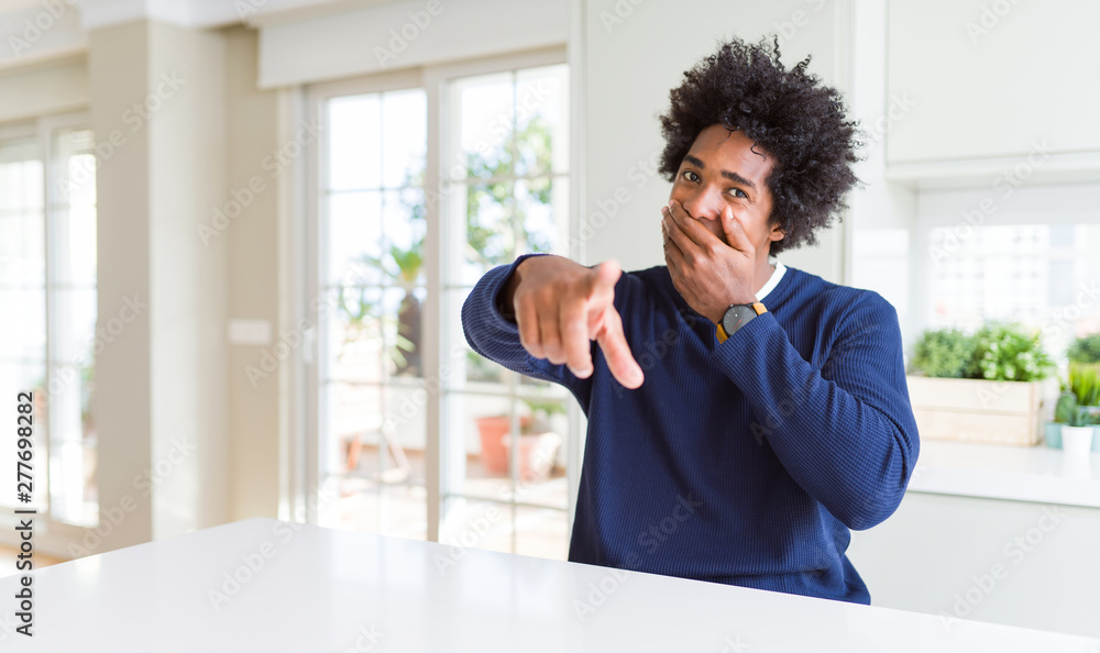Young african american man wearing casual sweater sitting at home laughing at you, pointing finger to the camera with hand over mouth, shame expression