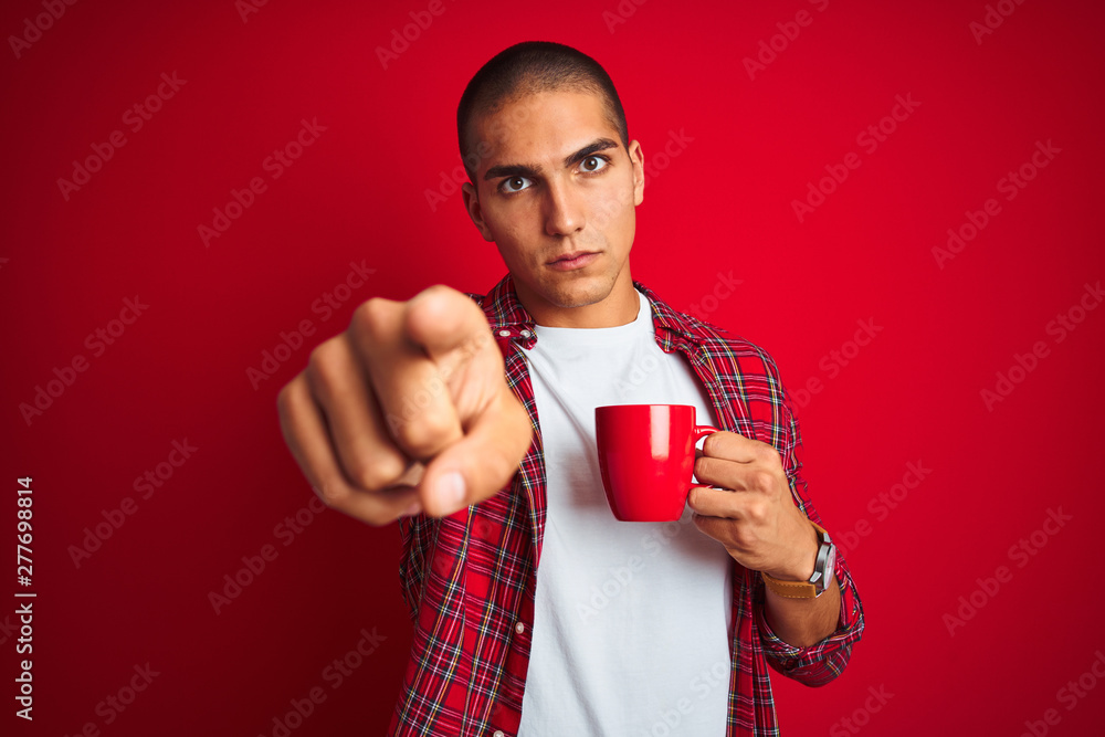 Young handsome man wearing shirt drinking a cup of coffee over red isolated background pointing with finger to the camera and to you, hand sign, positive and confident gesture from the front Young handsome man wearing shirt drinking a cup of coffee over red isolated background pointing with finger to the camera and to you, hand sign, positive and confident gesture from the front