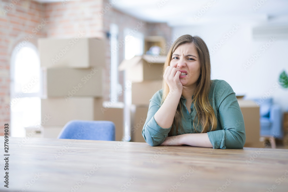 Young woman sitting on the table with cardboard boxes behind her moving to new home looking stressed and nervous with hands on mouth biting nails. Anxiety problem. Young woman sitting on the table with cardboard boxes behind her moving to new home looking stressed and nervous with hands on mouth biting nails. Anxiety problem.