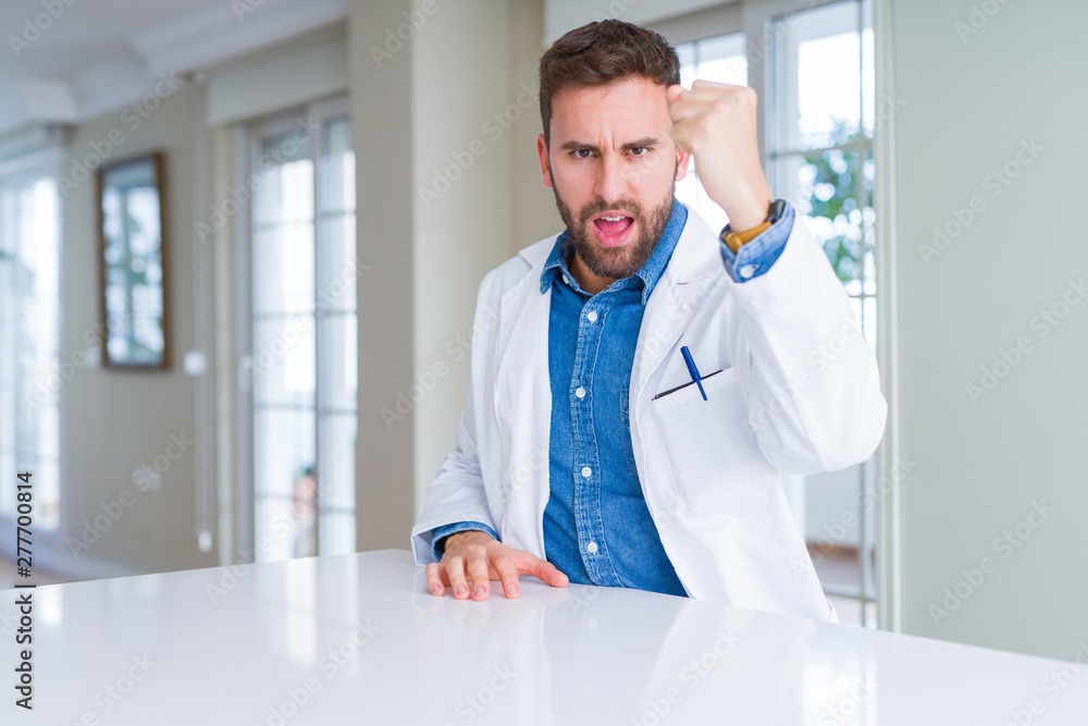 Handsome doctor man wearing medical coat at the clinic angry and mad raising fist frustrated and furious while shouting with anger. Rage and aggressive concept.