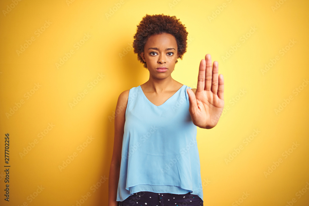 Beautiful african american woman wearing elegant shirt over isolated yellow background doing stop sing with palm of the hand. Warning expression with negative and serious gesture on the face. Beautiful african american woman wearing elegant shirt over isolated yellow background doing stop sing with palm of the hand. Warning expression with negative and serious gesture on the face.