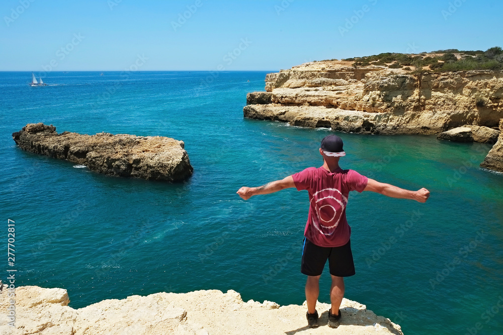Young man wearing tie dye t-shirt enjoying the panoramic top view of rocky beaches with cliffs somwhere, somwhere in Algarve, Portugal. Atlantic ocean shore background. Copy space for text.