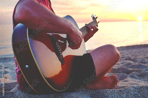 Young man wearing purple tie dye t-shirt playing dreadnought parlor acoustic guitar on beach at beautiful sunset time. Fit guitarist w/ sunburst instrument by the sea. Background, copy space, close up photo