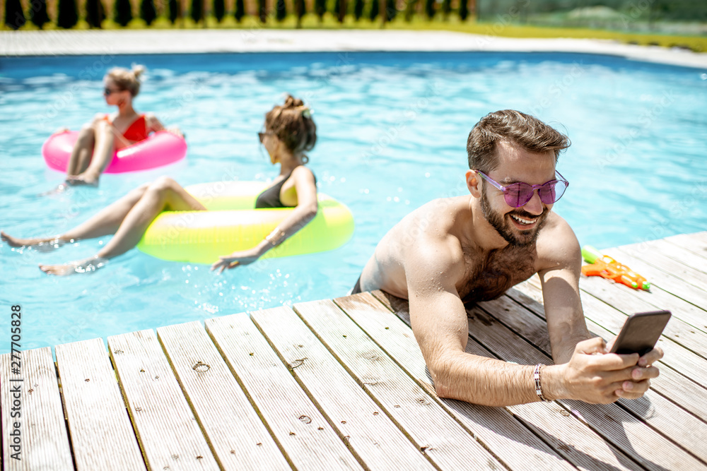 Man using smart phone while resting on the poolside during the summer vacations with friends in the swimming pool