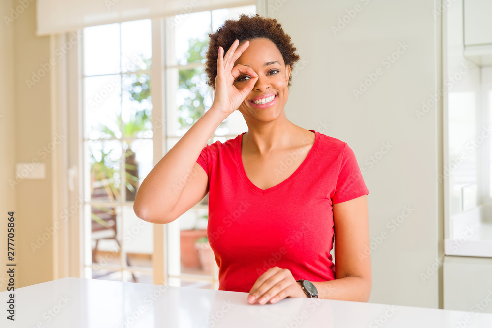Young beautiful african american woman at home doing ok gesture with hand smiling, eye looking through fingers with happy face. Young beautiful african american woman at home doing ok gesture with hand smiling, eye looking through fingers with happy face.