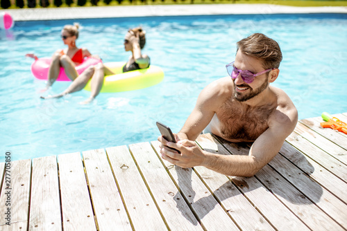 Man using smart phone while resting on the poolside during the summer vacations with friends in the swimming pool